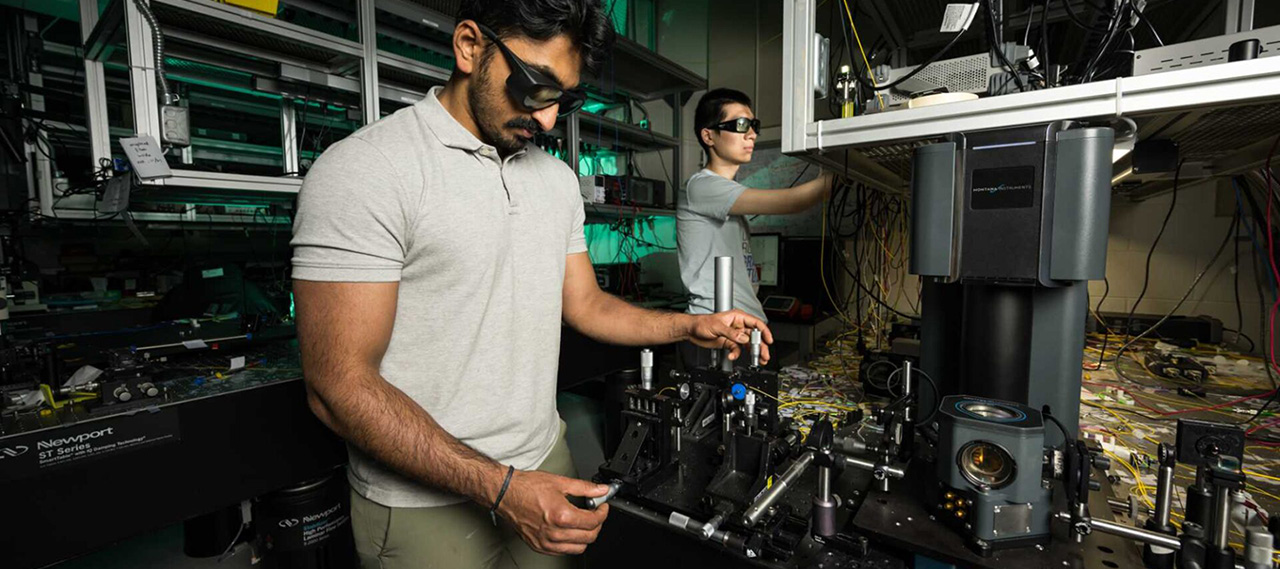 Two researchers wearing protective eyewear manipulate the equipment in an optics lab to produce surface acoustic waves.